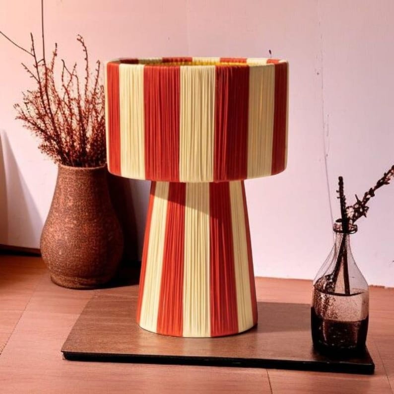 May include: A decorative table lamp with a cylindrical shade and base, featuring vertical stripes in red and cream. The lamp is made of yarn or thread, creating a textured surface. The lamp sits on a dark brown wooden base.