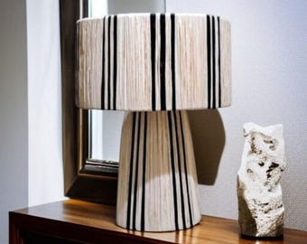 Può includere: Lampada da tavolo decorativa con paralume cilindrico e base conica. La lampada presenta un paralume e una base color crema con strisce nere verticali. Accanto alla lampada c'è una piccola scultura bianca strutturata.