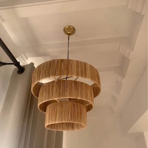 Puede incluir: Una lámpara de araña de tres niveles hecha de fibras naturales, con una montura de techo dorada. La lámpara tiene un diseño en capas, con cada nivel disminuyendo de tamaño. La lámpara cuelga de un cable negro. El fondo incluye un techo blanco y una cortina gris.