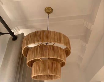 May include: A three-tiered, natural fiber chandelier with a gold-colored ceiling mount. The light fixture has a layered design, with each tier decreasing in size. The light fixture hangs from a black cord. The background includes a white ceiling and a gray curtain.