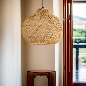 May include: A woven rattan pendant light. The light has a rounded shape with a woven pattern. It hangs from a black cord. The background includes a window and a small wooden table. The light is suspended from the ceiling.