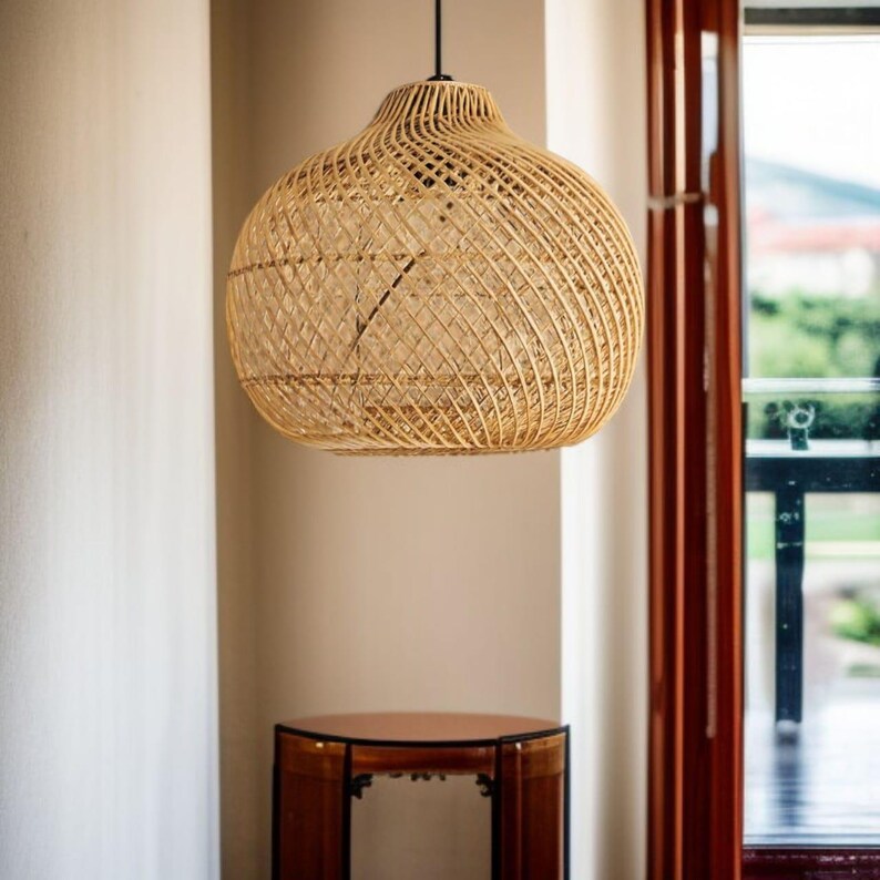 May include: A woven rattan pendant light. The light has a rounded shape with a woven pattern. It hangs from a black cord. The background includes a window and a small wooden table. The light is suspended from the ceiling.