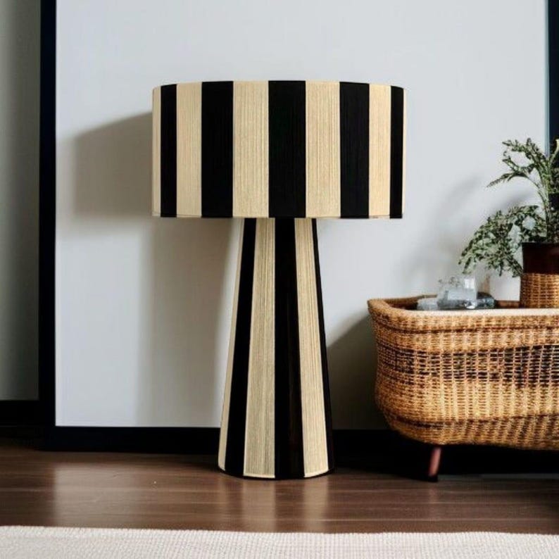 May include: A black and beige striped table lamp with a cylindrical lampshade and a matching base. The lamp is placed on a wooden floor, with a woven basket and a plant in the background.