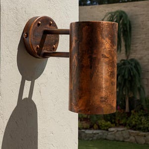May include: A copper-colored outdoor wall sconce. The cylindrical light fixture is mounted on a textured, off-white wall. The sconce has a circular backplate and a cylindrical shade, with a rustic, weathered finish. The sconce is mounted on a textured, off-white wall.