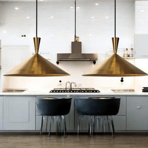 Może przedstawiać: Dwie mosiężne lampy wiszące w kształcie stożka wiszą nad wyspą kuchenną. Lampy mają złote wykończenie i czarne przewody. Poniżej dwa czarne krzesła z przezroczystymi nogami stoją przed białym blatem. Kuchnia posiada białe szafki i okap ze stali nierdzewnej.