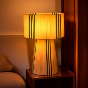 Puede incluir: L&aacute;mpara de mesa decorativa con pantalla cil&iacute;ndrica y base c&oacute;nica. La l&aacute;mpara est&aacute; hecha de un material claro, posiblemente tejido, con rayas verticales de color verde oscuro. La l&aacute;mpara est&aacute; encendida, emitiendo un brillo c&aacute;lido.