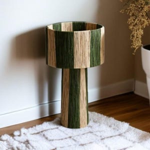 May include: A tall, decorative lamp with a cylindrical shade and base. The shade and base are wrapped in alternating vertical stripes of green and natural-colored woven material. The lamp sits on a white rug.