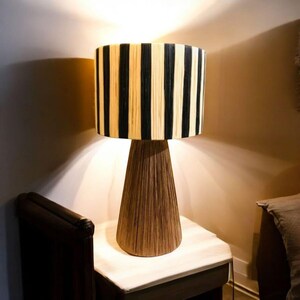 Op de afbeelding: Een decoratieve tafellamp met een cilindrische lampenkap met verticale zwarte en beige strepen. De lamp heeft een conische voet van bruin, getextureerd materiaal. De lamp staat op een kleine, lichtgekleurde tafel. Hoogte ongeveer 40 cm.