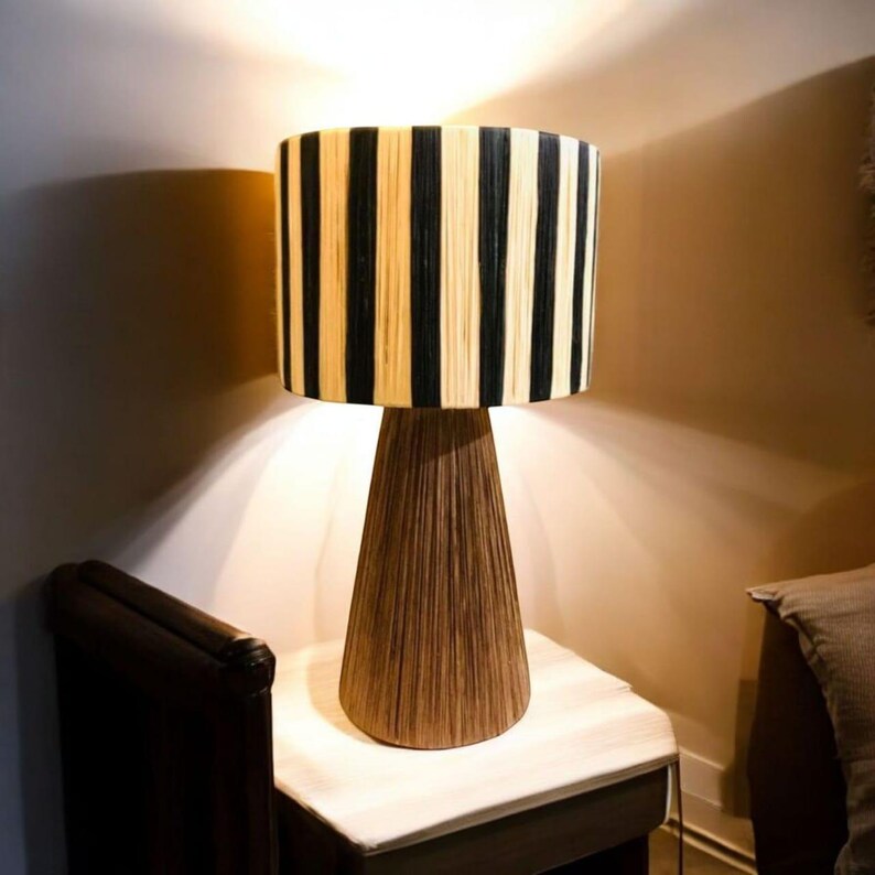 Op de afbeelding: Een decoratieve tafellamp met een cilindrische lampenkap met verticale zwarte en beige strepen. De lamp heeft een conische voet van bruin, getextureerd materiaal. De lamp staat op een kleine, lichtgekleurde tafel. Hoogte ongeveer 40 cm.