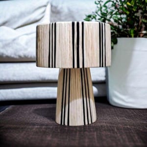 Puede incluir: Una l&aacute;mpara de mesa decorativa con una pantalla cil&iacute;ndrica y una base c&oacute;nica. La l&aacute;mpara presenta un color beige natural con rayas negras verticales. La l&aacute;mpara est&aacute; colocada sobre una superficie marr&oacute;n.
