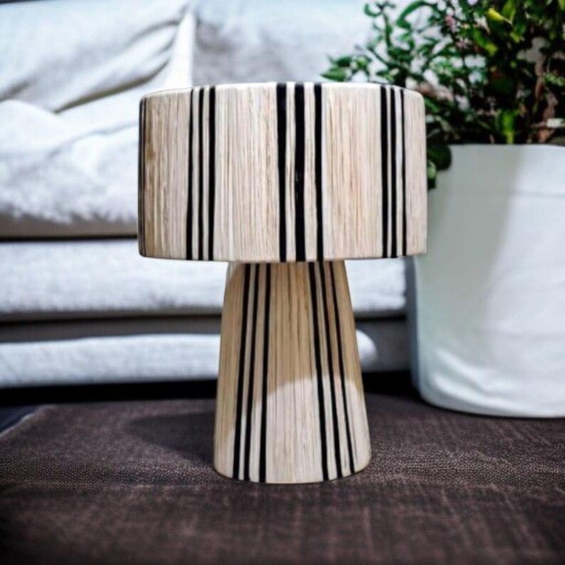 Puede incluir: Una l&aacute;mpara de mesa decorativa con una pantalla cil&iacute;ndrica y una base c&oacute;nica. La l&aacute;mpara presenta un color beige natural con rayas negras verticales. La l&aacute;mpara est&aacute; colocada sobre una superficie marr&oacute;n.