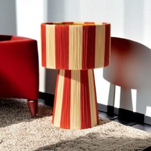 May include: A decorative table lamp with a red and cream striped pattern. The lamp has a cylindrical shade and a conical base, both adorned with vertical stripes. The lamp is made of a woven material and sits on a neutral-colored rug.