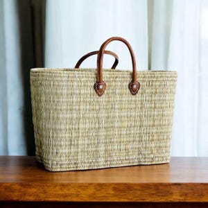 K&ouml;nnte beinhalten: Eine rechteckige Stroh-Tragetasche mit braunen Ledergriffen und Akzenten. Die Tasche ist hellbraun und steht auf einer Holzoberfl&auml;che. Die Griffe sind gew&ouml;lbt und mit Lederaufn&auml;hern befestigt.