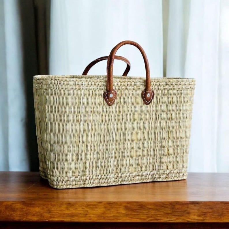 K&ouml;nnte beinhalten: Eine rechteckige Stroh-Tragetasche mit braunen Ledergriffen und Akzenten. Die Tasche ist hellbraun und steht auf einer Holzoberfl&auml;che. Die Griffe sind gew&ouml;lbt und mit Lederaufn&auml;hern befestigt.