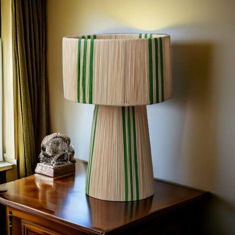 Puede incluir: L&aacute;mpara de mesa decorativa con una pantalla cil&iacute;ndrica y una base c&oacute;nica. La pantalla y la base son de color beige con rayas verdes verticales. La l&aacute;mpara est&aacute; colocada sobre una mesa de madera oscura, cerca de una ventana con cortinas verdes.