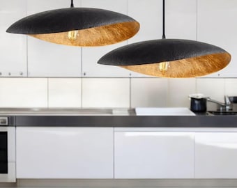 Op de afbeelding: Twee zwarte hanglampen met een gehamerde textuur en goudkleurige interieurs hangen in een keuken. De lampen hebben een druppelvorm en worden door zwarte snoeren aan het plafond gehangen. De keuken heeft witte kasten en een roestvrijstalen werkblad.