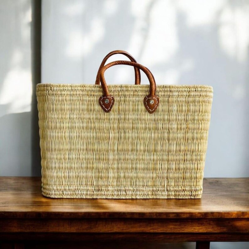 K&ouml;nnte beinhalten: Rechteckige Stroh-Tragetasche mit braunen Ledergriffen und -akzenten. Die Tasche ist hellbeige und steht auf einer Holzoberfl&auml;che. Die Griffe sind mit braunen Lederaufn&auml;hern befestigt.