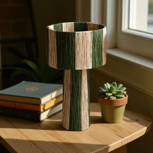 May include: A decorative table lamp with a cylindrical shade and base. The shade and base are wrapped in alternating vertical stripes of green and beige yarn. The lamp sits on a wooden table next to a small potted succulent and a stack of books.
