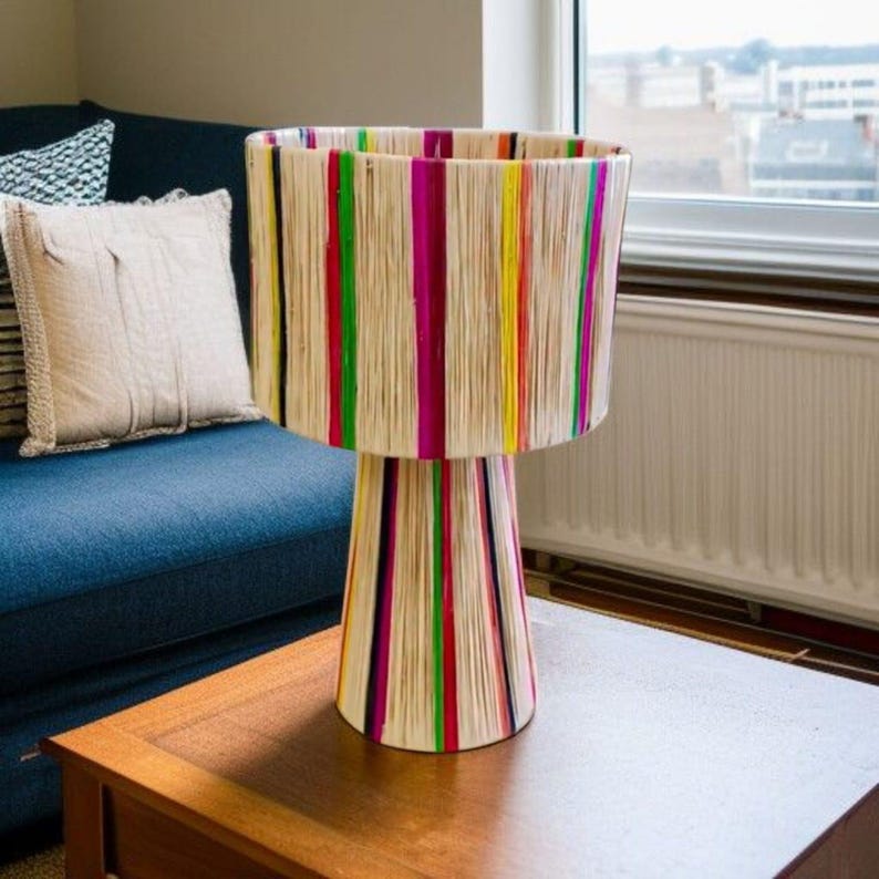 K&ouml;nnte beinhalten: Eine dekorative Tischlampe mit einem zylindrischen Schirm und einem konischen Fu&szlig;. Der Schirm ist mit vertikalen Streifen in verschiedenen Farben verziert, darunter Pink, Gr&uuml;n, Gelb und Rot. Die Lampe steht auf einem Holztisch.
