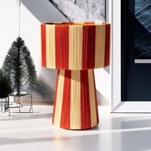 May include: A decorative lamp with a cylindrical shade and a pedestal base. The lamp features vertical stripes in alternating red and cream colors. The lamp is a unique home decor item.