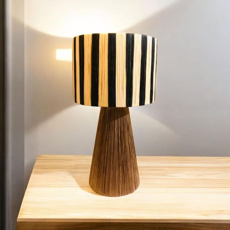 Op de afbeelding: Een decoratieve tafellamp met een cilindrische lampenkap met verticale zwarte en beige strepen. De lamp heeft een conische, bruine voet. De lamp staat op een houten tafel en cre&euml;ert een warme en uitnodigende sfeer. Hoogte: ongeveer 40 cm.