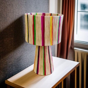 K&ouml;nnte beinhalten: Eine dekorative Tischlampe mit einem zylindrischen Lampenschirm und einem konischen Sockel. Der Lampenschirm und der Sockel sind mit vertikalen Streifen in verschiedenen Farben verziert, darunter Rosa, Gr&uuml;n, Gelb und Schwarz. Die Lampe steht auf einem hellen Holztisch.