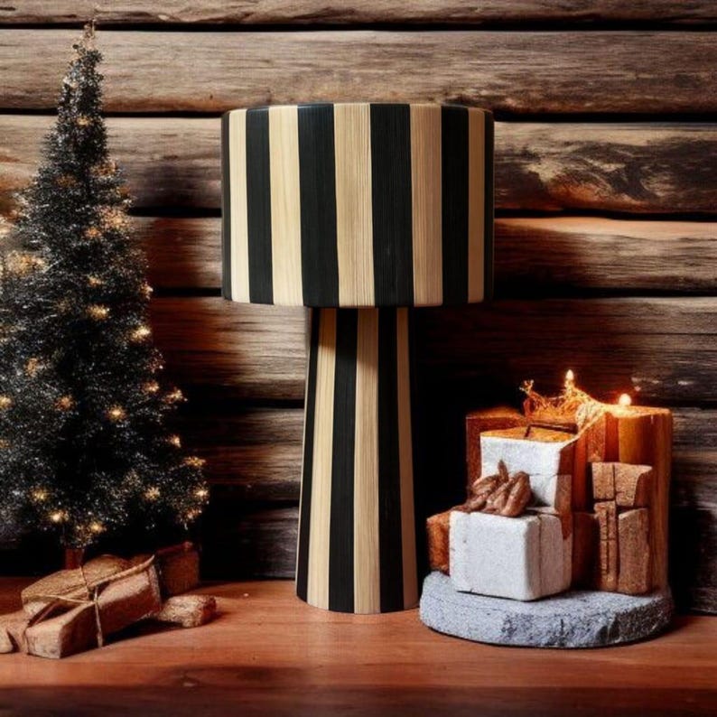 Op de afbeelding: Een decoratieve lamp met een zwart-beige gestreepte kap en voet. Links staat een kleine zwarte kerstboom met lichtjes. Er zijn verschillende ingepakte cadeaus en kaarsen.