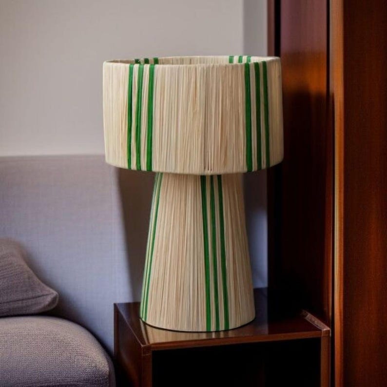 Puede incluir: Una l&aacute;mpara de mesa decorativa con una pantalla cil&iacute;ndrica y una base c&oacute;nica. La l&aacute;mpara est&aacute; hecha de un material de color natural con rayas verdes verticales. Se encuentra sobre una mesita de noche de madera oscura.