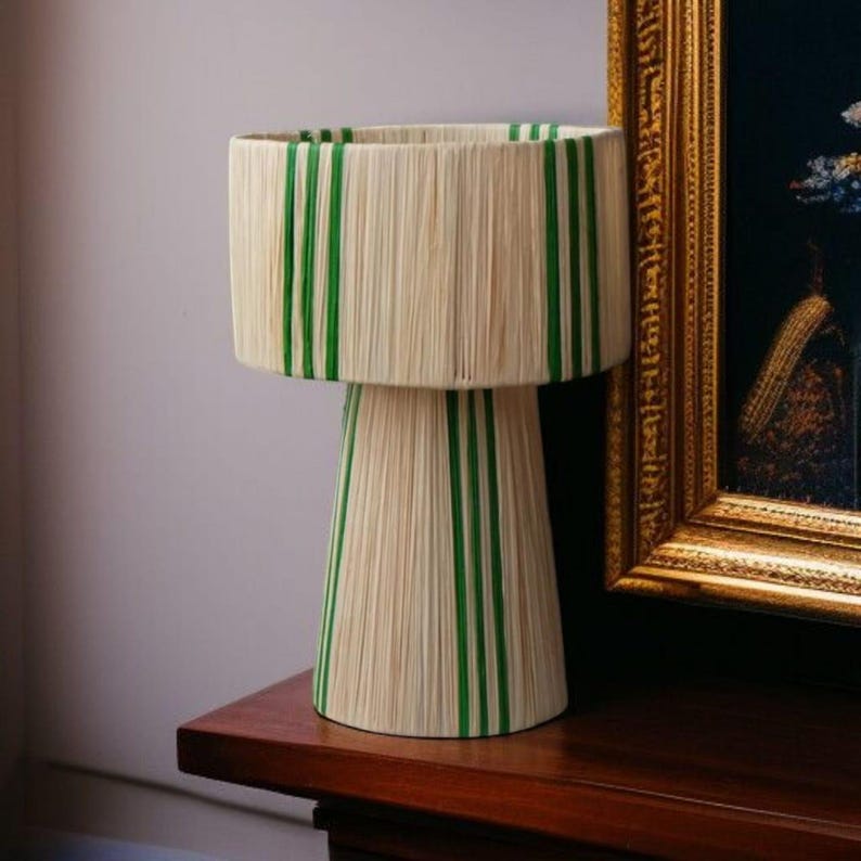 Puede incluir: Una l&aacute;mpara de mesa decorativa con pantalla y base cil&iacute;ndricas. La l&aacute;mpara presenta un material tejido blanco roto natural con rayas verdes verticales. La l&aacute;mpara est&aacute; colocada sobre una superficie de madera oscura.