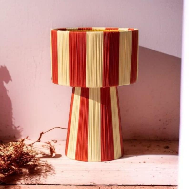 May include: A decorative table lamp with a cylindrical lampshade and a conical base. The lamp is adorned with vertical stripes of red and cream-colored yarn, creating a visually striking design. The lamp is a unique home decor item.
