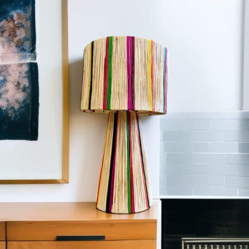 K&ouml;nnte beinhalten: Eine dekorative Tischlampe mit zylindrischem Lampenschirm und konischem Fu&szlig;. Die Lampe besteht aus naturfarbenem Material mit vertikalen Streifen in Pink, Gr&uuml;n, Gelb und Schwarz. Die Lampe steht auf einer Holzoberfl&auml;che.