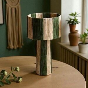 May include: A decorative lamp with a cylindrical shade and base, featuring alternating vertical stripes of green and natural beige. The lamp is made of woven material and sits on a round wooden table. The overall design is modern and natural.