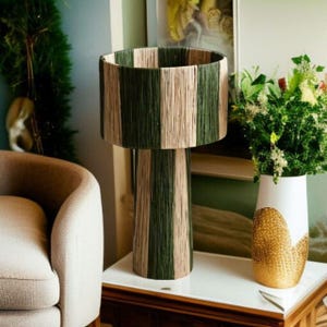 May include: A decorative lamp with a cylindrical shade and base, wrapped in alternating vertical stripes of green and beige yarn. The lamp sits on a white table, next to a white vase with a gold textured pattern and a bouquet of flowers.