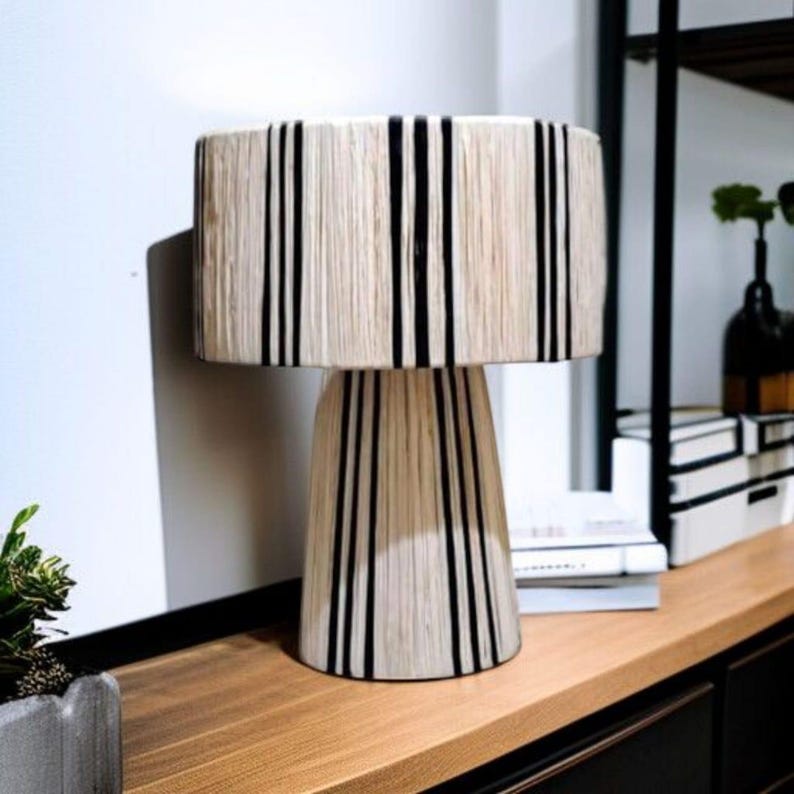 Puede incluir: Una l&aacute;mpara de mesa decorativa con una pantalla cil&iacute;ndrica y una base c&oacute;nica. La l&aacute;mpara presenta un color beige natural con rayas negras verticales. La l&aacute;mpara est&aacute; colocada sobre una superficie de madera, a&ntilde;adiendo un toque de dise&ntilde;o moderno a cualquier habitaci&oacute;n.