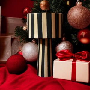 Op de afbeelding: Een decoratieve lamp met een zwart-beige streepjespatroon, geplaatst tegen een kerstboom. De sc&egrave;ne bevat rode en zilveren ornamenten, een ingepakt cadeau met een rode strik en een rood oppervlak.