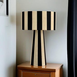 May include: A black and beige striped table lamp. The lamp has a cylindrical lampshade with vertical black and beige stripes. The base of the lamp is a matching black and beige striped design. The lamp is sitting on a wooden surface.