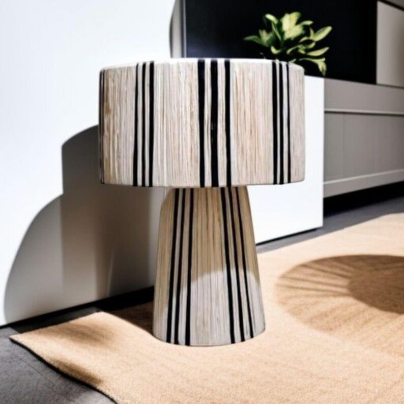 Puede incluir: Una l&aacute;mpara de mesa decorativa con una pantalla cil&iacute;ndrica y una base c&oacute;nica. La l&aacute;mpara presenta un material natural blanquecino con rayas negras verticales. La l&aacute;mpara est&aacute; colocada sobre una alfombra de color neutro.