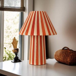 Könnte beinhalten: Eine dekorative Tischlampe mit konischem Schirm und zylindrischem Sockel. Die Lampe hat vertikale Streifen in abwechselnden Orange- und Cremetönen. Die Lampe steht auf einer weißen Oberfläche.