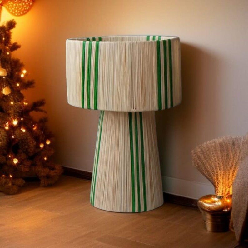 Puede incluir: Una l&aacute;mpara decorativa con pantalla y base cil&iacute;ndricas. La l&aacute;mpara est&aacute; envuelta en una fibra natural con rayas verdes verticales. La l&aacute;mpara est&aacute; sobre un suelo de madera, con un &aacute;rbol de Navidad al fondo.