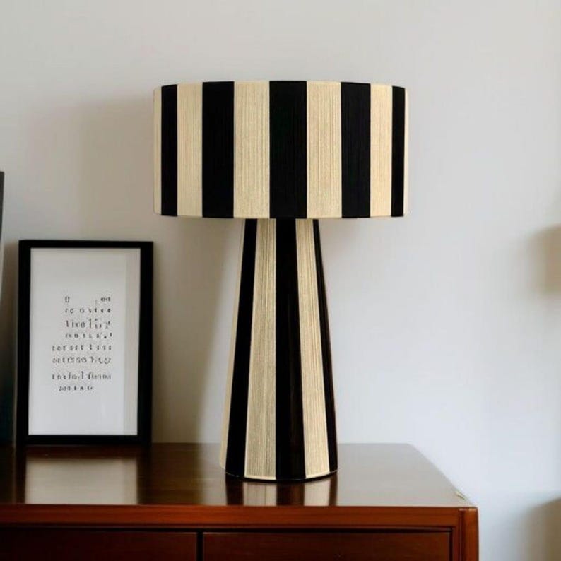 May include: A decorative table lamp with a black and cream striped shade and base. The lamp sits on a dark wood surface. A framed print with text is visible in the background. The lamp's design is modern and stylish.