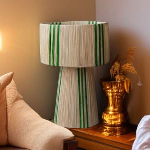 Puede incluir: Una l&aacute;mpara de mesa decorativa con pantalla y base cil&iacute;ndricas. La l&aacute;mpara presenta una textura tejida de color crema con rayas verdes verticales. La l&aacute;mpara est&aacute; junto a un jarr&oacute;n dorado con plantas secas.