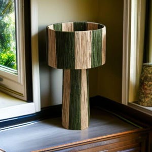May include: A decorative lamp with a cylindrical shade and base. The shade and base are wrapped in alternating vertical stripes of green and beige material. The lamp is placed on a gray surface.