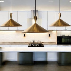 K&ouml;nnte beinhalten: Drei moderne Pendelleuchten mit konischen Lampenschirmen in Messingoptik h&auml;ngen &uuml;ber einer wei&szlig;en K&uuml;cheninsel. Die Leuchten haben ein schlankes, minimalistisches Design mit einem schmalen, zylindrischen Oberteil und einem breiten, nach unten gerichteten Kegel. Die K&uuml;che verf&uuml;gt &uuml;ber wei&szlig;e Schr&auml;nke und eine hellfarbige Arbeitsplatte.