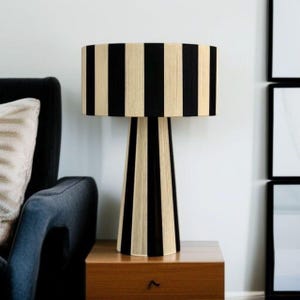 May include: A black and beige striped table lamp with a cylindrical lampshade and a conical base. The lamp is placed on a wooden nightstand next to a dark gray armchair. The lamp's height is approximately 24 inches.