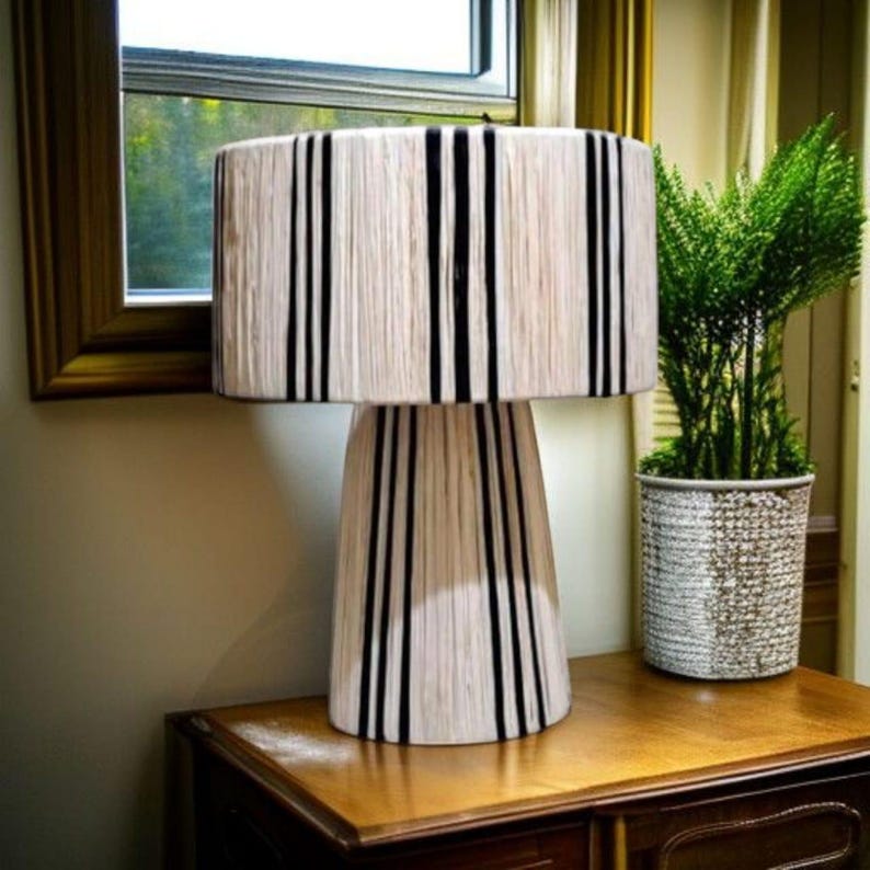 Puede incluir: Una l&aacute;mpara de mesa decorativa con pantalla y base color crema, acentuada por rayas negras verticales. La l&aacute;mpara est&aacute; sobre una superficie de madera, junto a una planta en maceta. El dise&ntilde;o es moderno y adecuado para la decoraci&oacute;n del hogar.