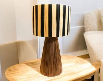 Può includere: Lampada da tavolo decorativa con paralume cilindrico a strisce verticali nere e beige. La lampada ha una base conica marrone e poggia su un tavolo di legno chiaro. Un elegante elemento di arredo per la casa.