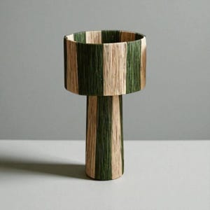 May include: A decorative lamp with a cylindrical shade and base. The shade and base are adorned with vertical stripes of green and beige woven material. The lamp has a modern design and is suitable for home decor.