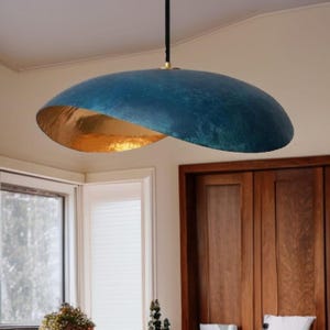Op de afbeelding: Een moderne hanglamp met een blauwe buitenkant en een gouden binnenkant. Het licht hangt aan een zwart snoer en heeft een messingkleurige accent. De armatuur heeft een unieke, gebogen vorm en voegt een stijlvolle touch toe aan elke kamer.