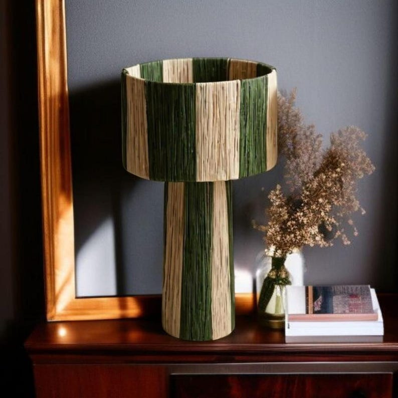 May include: A decorative table lamp with a cylindrical lampshade and base. The lampshade and base are vertically striped in alternating shades of green and beige. The lamp sits on a dark wooden surface, with a framed picture and a vase of dried flowers nearby.