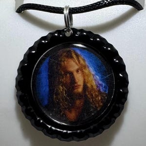 May include: Black bottle cap pendant necklace featuring a portrait of a person with long, wavy blonde hair against a blue background. The pendant is attached to a black cord necklace.